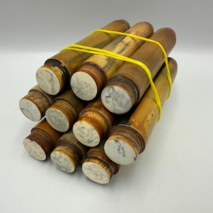 May include: A bundle of cylindrical wooden objects in light brown and tan hues, held together by a yellow elastic band. The ends of the cylinders feature a white and grey marbled pattern.