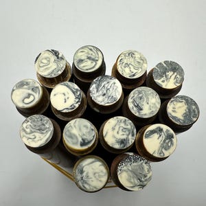 May include: A collection of twelve cylindrical wooden objects arranged in a cluster. Each object features a flat, circular top surface with a distinct black and white marbled pattern, while the bodies are made of a smooth, dark brown wood.