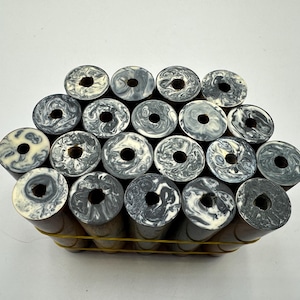 May include: A collection of nineteen cylindrical rods bundled together with a yellow rubber band. Each rod features a dark brown wooden body and a circular top end with a marbled pattern of black, grey, and white swirls and a central hole.