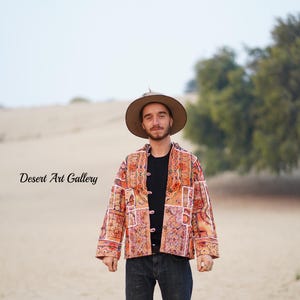 May include: A person wearing a patterned jacket with orange, red, and white accents, black shirt, blue jeans, and a brown hat. The text "Desert Art Gallery" is visible in the image.