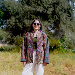 May include: A woman wearing a colorful patchwork jacket with a green top and white pants. The jacket features intricate patterns in shades of brown, purple, and teal. The image is taken outdoors with the text "Desert Art Gallery" in the top right corner.