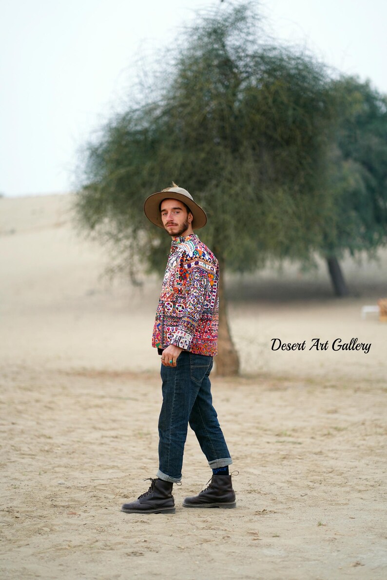 Op de afbeelding: Een persoon met een overhemd met patroon, een blauwe spijkerbroek en een bruine hoed staat in een woestijnlandschap. Het overhemd heeft een kleurrijk, ingewikkeld ontwerp. De tekst "Desert Art Gallery" is zichtbaar in de afbeelding.