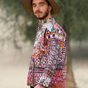 Op de afbeelding: Een kleurrijk, patroonshirt met een knoopsluiting. Het shirt heeft een levendige mix van oranje, roze, blauwe en witte geometrische patronen. De persoon draagt een bruine hoed en een blauwe spijkerbroek. Het shirt is een uniek kledingstuk.