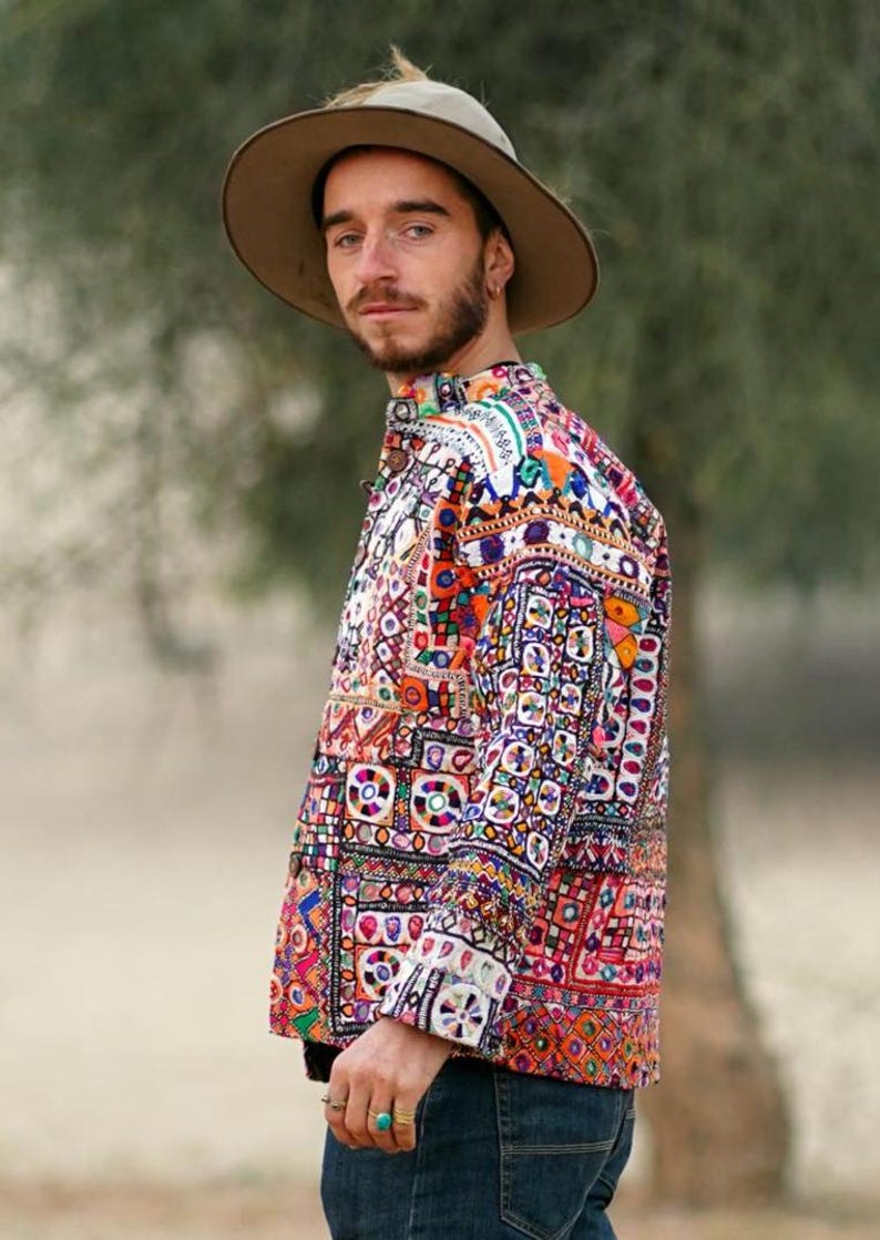 Op de afbeelding: Een kleurrijk, patroonshirt met een knoopsluiting. Het shirt heeft een levendige mix van oranje, roze, blauwe en witte geometrische patronen. De persoon draagt een bruine hoed en een blauwe spijkerbroek. Het shirt is een uniek kledingstuk.