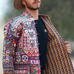 Op de afbeelding: Een kleurrijke, geborduurde jas met een bruine hoed. De jas heeft een patchwork-ontwerp met verschillende geometrische patronen en levendige kleuren, waaronder rood, blauw en oranje. De binnenvoering heeft een geel en zwart zigzagpatroon.
