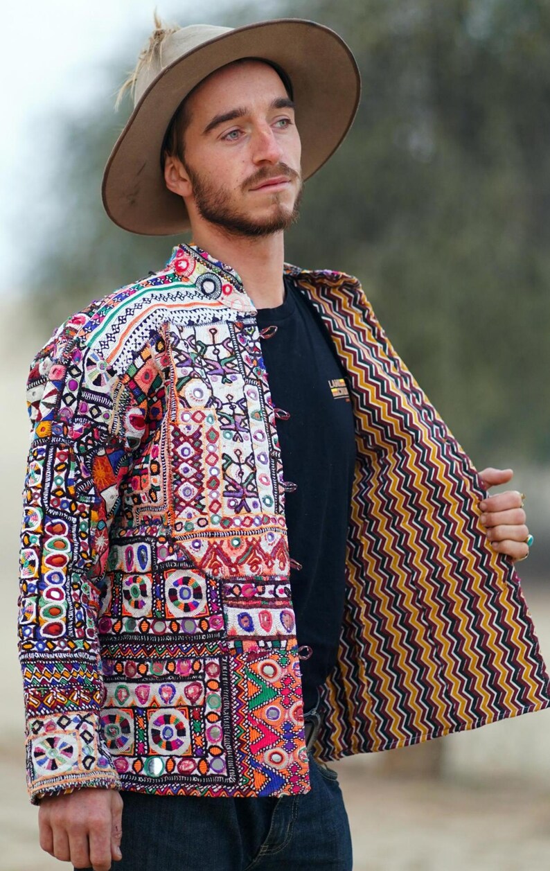 Op de afbeelding: Een kleurrijke, geborduurde jas met een bruine hoed. De jas heeft een patchwork-ontwerp met verschillende geometrische patronen en levendige kleuren, waaronder rood, blauw en oranje. De binnenvoering heeft een geel en zwart zigzagpatroon.