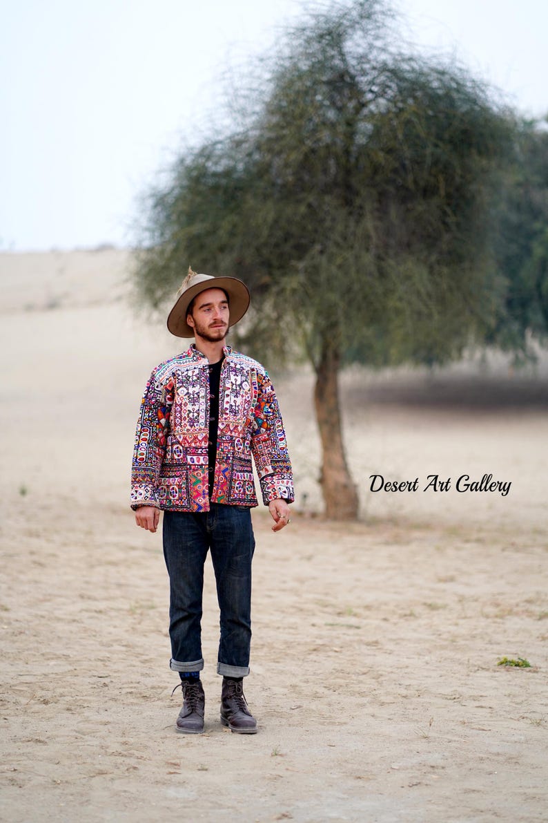 Op de afbeelding: Een kleurrijke jas met patronen met een verscheidenheid aan geometrische en bloemmotieven. De jas wordt gedragen met een donkerblauwe jeans en bruine laarzen. Een bruine hoed wordt gedragen. De tekst "Desert Art Gallery" is zichtbaar.