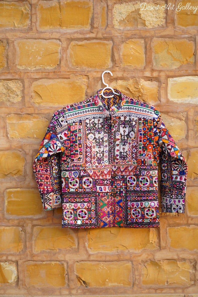 Op de afbeelding: Een kleurrijke jas met lange mouwen, ingewikkelde borduursels en spiegelaccenten. De jas heeft een knoopsluiting en een levendig patchwork-ontwerp in tinten rood, oranje, groen en blauw. De tekst "Desert Art Gallery" is rechtsboven zichtbaar.