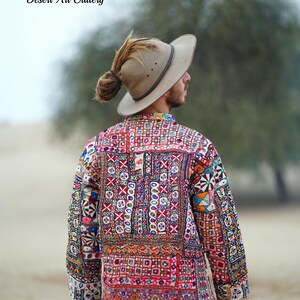 Op de afbeelding: Een kleurrijke, geborduurde jas met een patchwork ontwerp. De jas heeft verschillende geometrische patronen en levendige kleuren, waaronder rood, blauw en groen. De tekst "Desert Art Gallery" is zichtbaar. De persoon draagt een beige hoed.