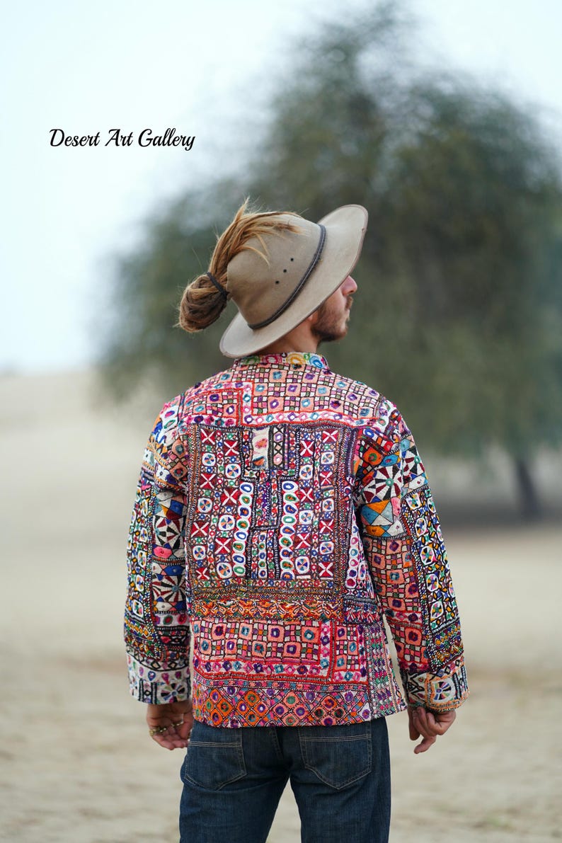 Op de afbeelding: Een kleurrijke, geborduurde jas met een patchwork ontwerp. De jas heeft verschillende geometrische patronen en levendige kleuren, waaronder rood, blauw en groen. De tekst "Desert Art Gallery" is zichtbaar. De persoon draagt een beige hoed.
