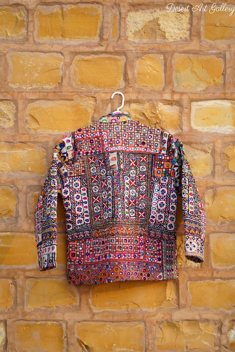 Op de afbeelding: Een kleurrijke, ingewikkeld geborduurde jas met een geometrisch patroon, hangend aan een witte hanger tegen een bakstenen muur. De jas heeft verschillende kleuren, waaronder rood, blauw en paars, en is versierd met kleine spiegels.