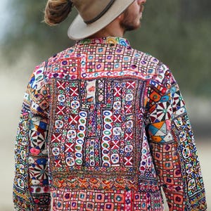 Op de afbeelding: Een kleurrijke jas met ingewikkelde borduursels in rood, oranje, blauw en groen. Het ontwerp bevat geometrische vormen en kruissteekpatronen, waardoor een levendig en gedetailleerd textiel ontstaat. De jas wordt gedragen met een beige hoed.