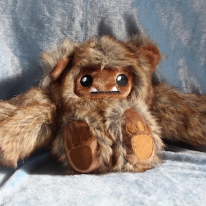 May include: A plush toy in the shape of a brown furry creature with large eyes, small ears, and a rectangular face. The toy has a light brown belly and paw pads. The background is a light blue fabric.