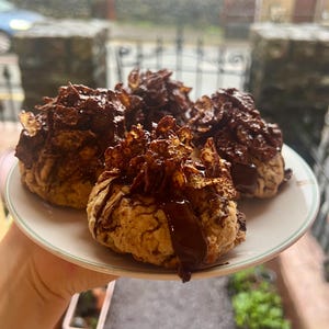 May include: A plate holds four baked goods, each topped with a dark chocolate coating and crispy cereal. The treats are light brown with a drizzle of chocolate. The plate has a white surface with a green rim. The background is blurred.