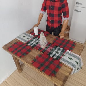 May include: A wooden table with a red and green plaid table runner and a white table runner with a green tree design. There is a small white cup and a small white artificial tree on the table.
