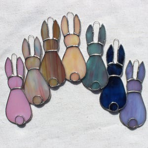 May include: A collection of seven stained glass bunny ornaments in various colors, including pink, yellow, blue, and purple. Each bunny has a silver-colored metal outline and a small circular tail. The ornaments are arranged on a white surface.