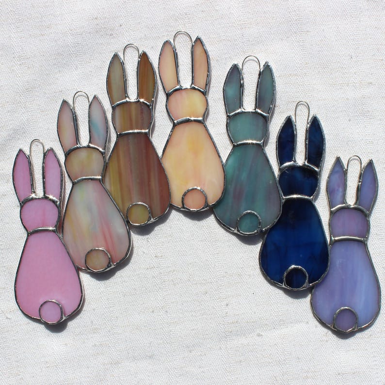 May include: A collection of seven stained glass bunny ornaments in various colors, including pink, yellow, blue, and purple. Each bunny has a silver-colored metal outline and a small circular tail. The ornaments are arranged on a white surface.