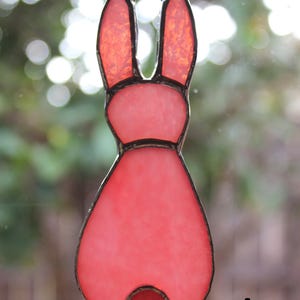 May include: A stained glass bunny suncatcher in shades of pink and red. The bunny has long ears, a round body, and a small tail. The glass is outlined with a dark border and is hanging from a black cord. The letter "A" is in the bottom right corner.