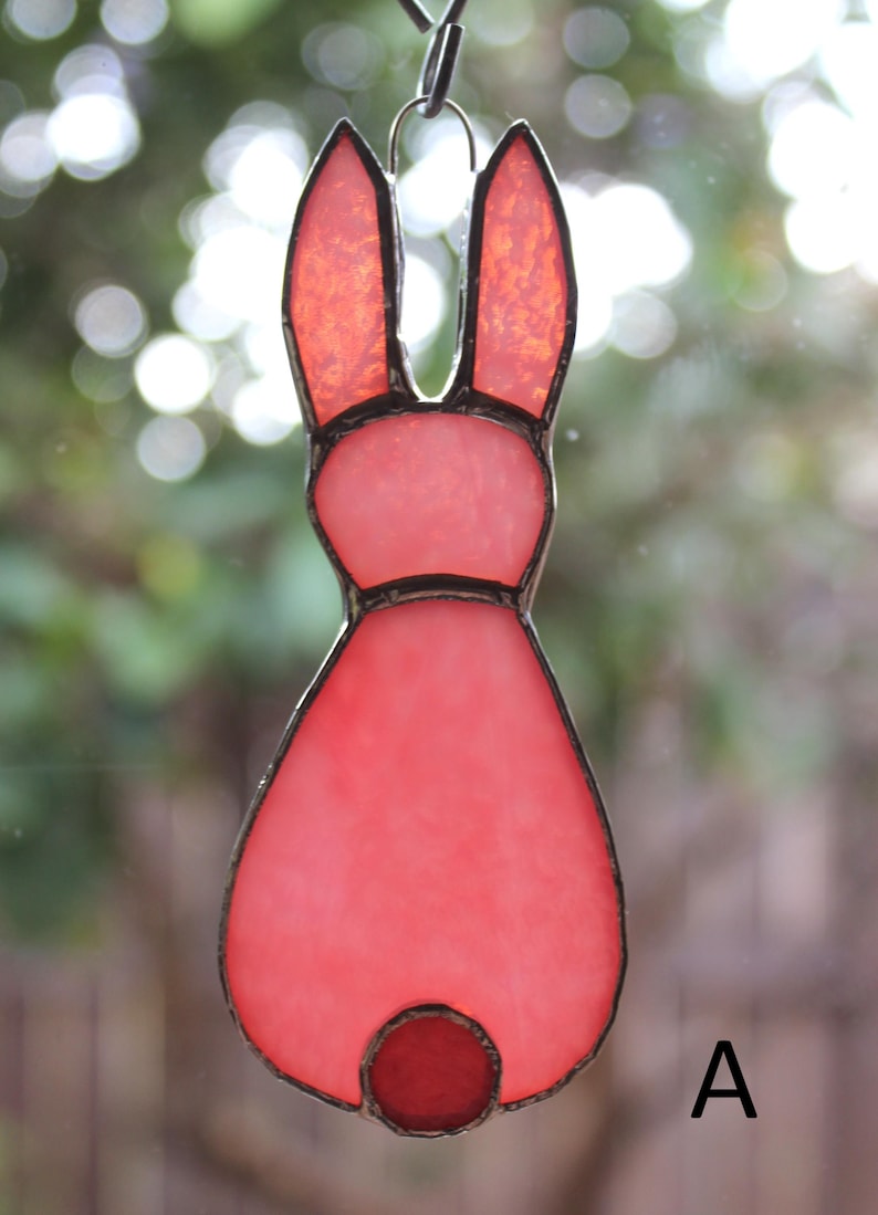 May include: A stained glass bunny suncatcher in shades of pink and red. The bunny has long ears, a round body, and a small tail. The glass is outlined with a dark border and is hanging from a black cord. The letter "A" is in the bottom right corner.