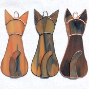 May include: Three stained glass cat ornaments in shades of orange, brown, and black. Each cat is depicted from the back, with pointed ears and a curled tail. The glass pieces are outlined with copper-coloured metal.