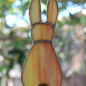 May include: Stained glass bunny suncatcher with a warm color palette of yellow, orange, and brown. The bunny has long ears and a round tail. The glass is outlined with a silver-colored metal. A decorative item for Easter or spring.
