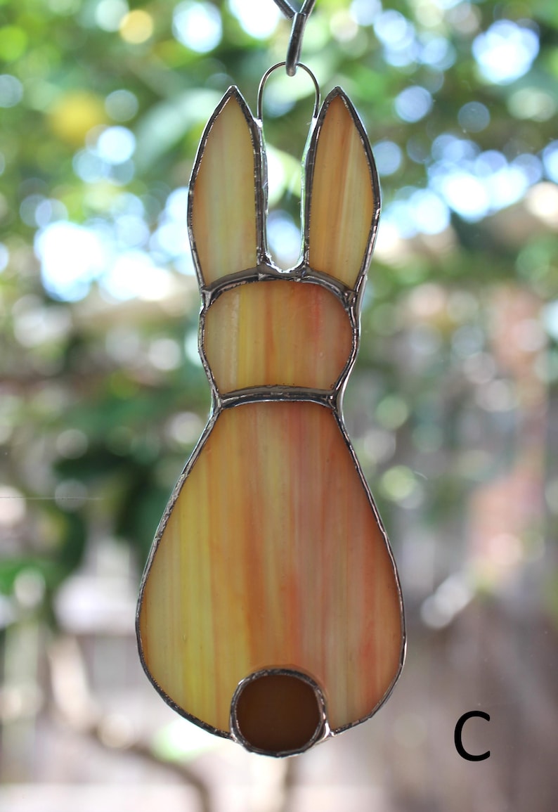May include: Stained glass bunny suncatcher with a warm color palette of yellow, orange, and brown. The bunny has long ears and a round tail. The glass is outlined with a silver-colored metal. A decorative item for Easter or spring.