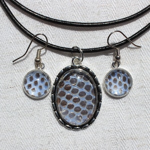 Peut inclure: Un collier et des boucles d'oreilles en argent avec un cordon en cuir noir et des pendentifs ovales et ronds avec un motif.