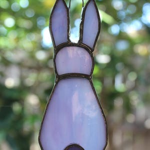 May include: A stained glass bunny ornament in shades of lavender and white, with a dark purple tail. The bunny is outlined in a dark metal frame and hangs from a silver chain. The background is a blurred green.
