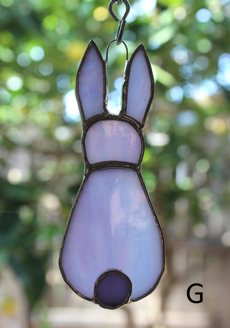May include: A stained glass bunny ornament in shades of lavender and white, with a dark purple tail. The bunny is outlined in a dark metal frame and hangs from a silver chain. The background is a blurred green.
