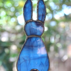 May include: A stained glass bunny ornament in shades of blue. The bunny has long ears and a round tail. The glass is framed with a silver-colored metal. A small metal loop is at the top for hanging.
