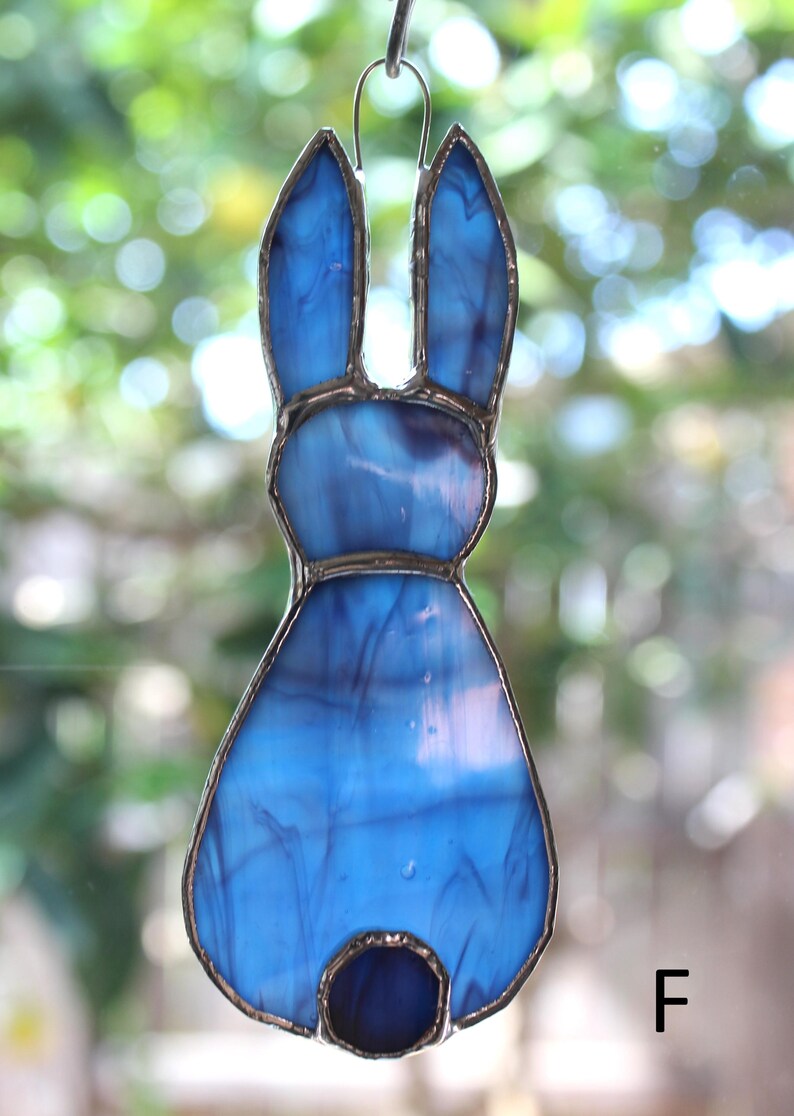 May include: A stained glass bunny ornament in shades of blue. The bunny has long ears and a round tail. The glass is framed with a silver-colored metal. A small metal loop is at the top for hanging.
