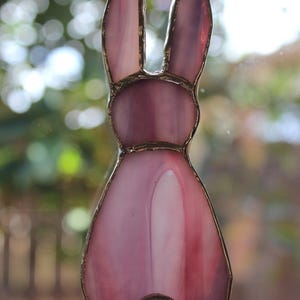 May include: A stained glass bunny decoration in shades of pink and purple. The bunny has long ears and a round tail. The glass is outlined with a gold-colored metal. The bunny is hanging from a metal loop.