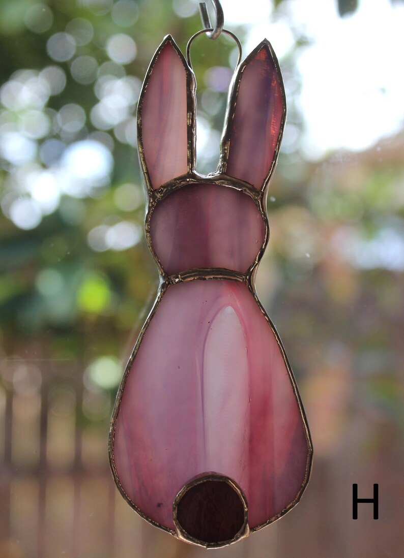 May include: A stained glass bunny decoration in shades of pink and purple. The bunny has long ears and a round tail. The glass is outlined with a gold-colored metal. The bunny is hanging from a metal loop.