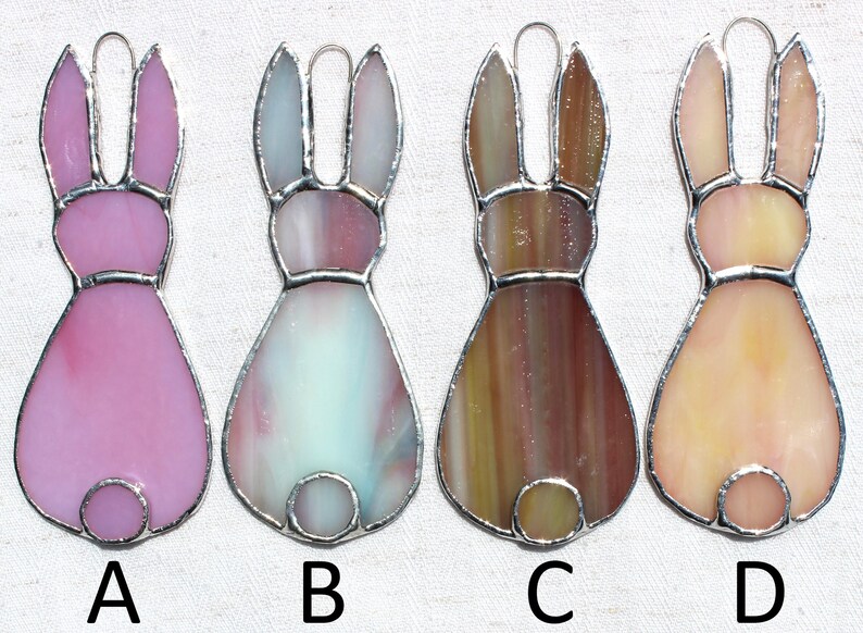 May include: Four stained glass bunny ornaments in shades of pink, blue, brown, and peach. Each bunny has a silver-colored metal outline and a loop for hanging. The ornaments are labeled A, B, C, and D.