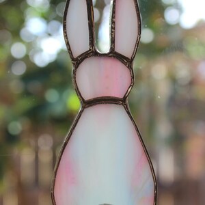 May include: A stained glass bunny suncatcher with a metal frame. The bunny features white, pink, and blue hues. The bunny's ears are white, and the body transitions from pink to white. A small, round, white bead forms the tail. The letter "B" is in the lower right corner.