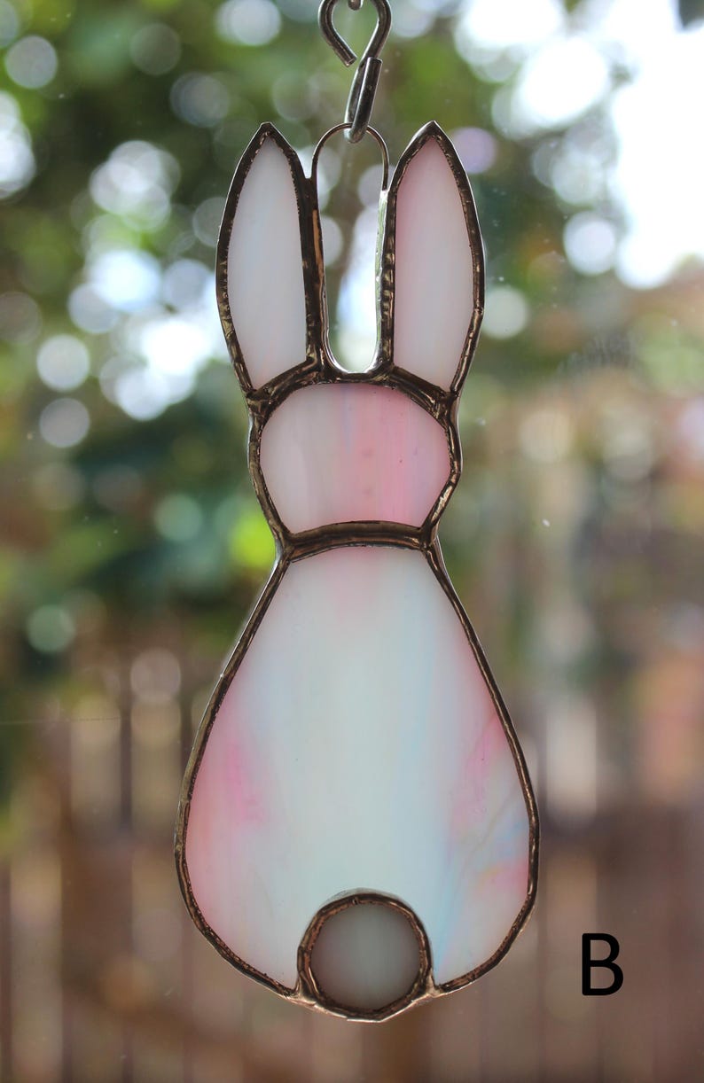 May include: A stained glass bunny suncatcher with a metal frame. The bunny features white, pink, and blue hues. The bunny's ears are white, and the body transitions from pink to white. A small, round, white bead forms the tail. The letter "B" is in the lower right corner.
