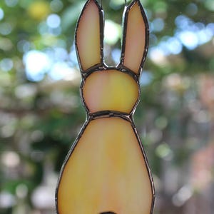May include: A stained glass bunny ornament with a yellow and orange gradient. The bunny has long ears and a small, round tail. The edges are outlined in a dark metal. A small metal loop is at the top for hanging.