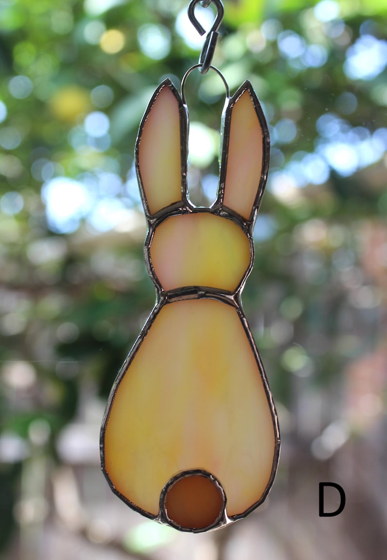 May include: A stained glass bunny ornament with a yellow and orange gradient. The bunny has long ears and a small, round tail. The edges are outlined in a dark metal. A small metal loop is at the top for hanging.