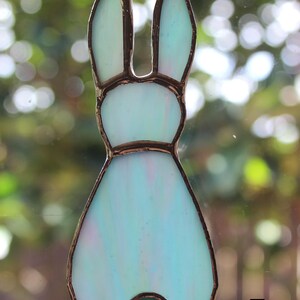 May include: A stained glass bunny suncatcher in shades of light blue and green, with a copper-colored border. The bunny has long ears and a round tail. The letter "E" is visible in the bottom right corner.