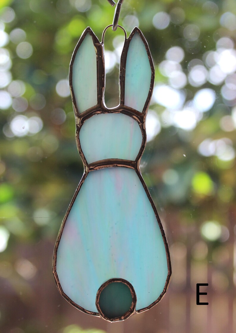 May include: A stained glass bunny suncatcher in shades of light blue and green, with a copper-colored border. The bunny has long ears and a round tail. The letter "E" is visible in the bottom right corner.