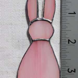 May include: A pink stained glass bunny ornament, approximately 3 inches tall, with a silver-colored metal border and a small loop for hanging. The bunny's body is a soft pink, with a lighter pink hue in the ears and tail.