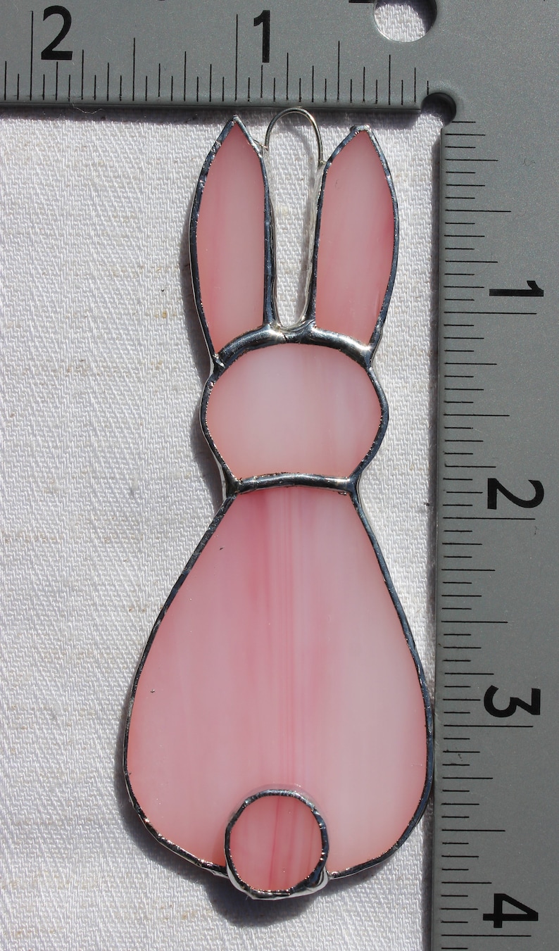May include: A pink stained glass bunny ornament, approximately 3 inches tall, with a silver-colored metal border and a small loop for hanging. The bunny's body is a soft pink, with a lighter pink hue in the ears and tail.