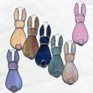 May include: A collection of stained glass bunny ornaments in various colors, including pink, blue, and yellow. Each bunny has a silver-colored metal outline and a small loop for hanging. The glass has a textured, translucent appearance.