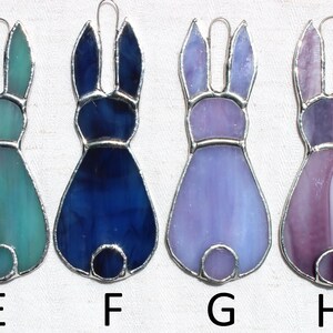 May include: Four stained glass bunny ornaments in shades of teal, blue, purple, and pink. Each bunny has a silver-colored metal outline and a loop for hanging. The letters E, F, G, and H are below each bunny.