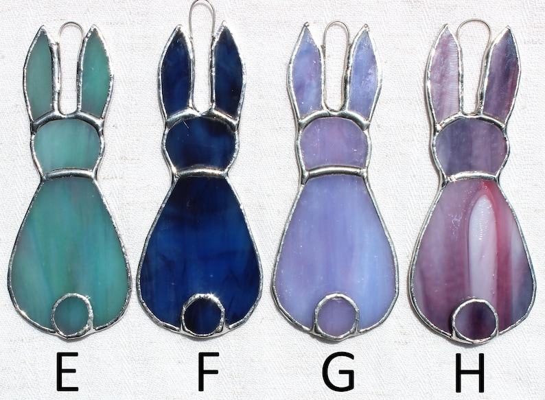 May include: Four stained glass bunny ornaments in shades of teal, blue, purple, and pink. Each bunny has a silver-colored metal outline and a loop for hanging. The letters E, F, G, and H are below each bunny.