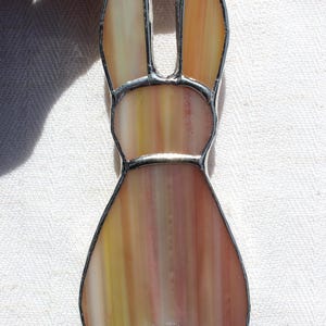 May include: A stained glass bunny ornament with a silver-colored metal border. The bunny is primarily shades of yellow, orange, and pink. The bunny has long ears, a round body, and a small, round tail. The letter "C" is visible in the lower right corner.