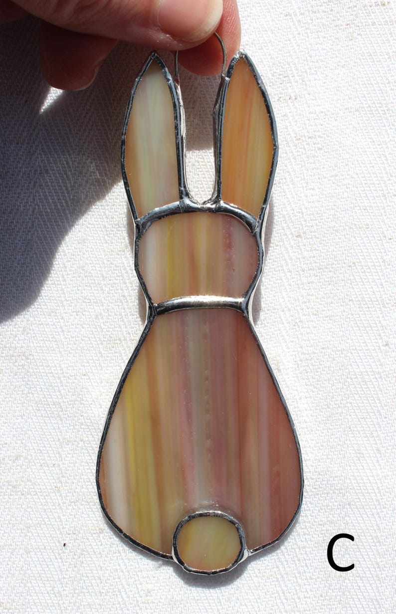 May include: A stained glass bunny ornament with a silver-colored metal border. The bunny is primarily shades of yellow, orange, and pink. The bunny has long ears, a round body, and a small, round tail. The letter "C" is visible in the lower right corner.