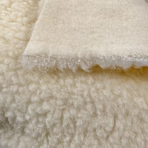 Könnte beinhalten: Nahaufnahme eines cremefarbenen Stoffes mit einer strukturierten, flauschigen Oberfläche. Ein Teil des Stoffes ist umgefaltet und zeigt eine glatte, flache Unterseite. Das Material wirkt weich und warm, geeignet für verschiedene Textilprojekte.