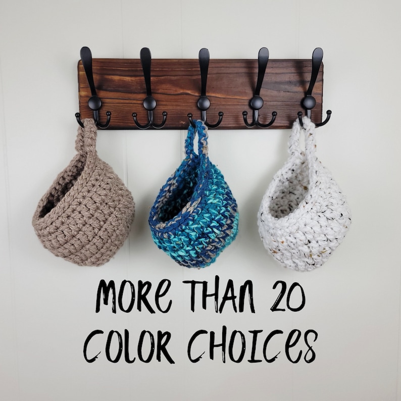 May include: Three hanging crocheted baskets in brown, blue and white, hanging on a wooden rack with black hooks. The text "MORE THAN 20 COLOR CHOICES" is below the baskets.