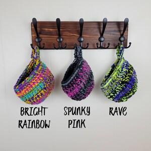 May include: Three crocheted hanging baskets in bright colours. The baskets are labelled "Bright Rainbow", "Spunky Pink", and "Rave".